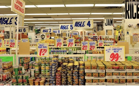 Crowded grocery display with handwritten promotional signs, varied price formats, and mixed product arrangements